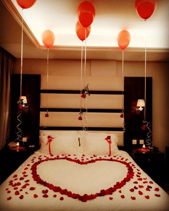 اتاق عروسی که تخت آن با گلبرگ های گل رز قرمز به شکل قلب تزیین شده است و از بادکنک های هلیومی شاخه های گل رز آویزان شده است